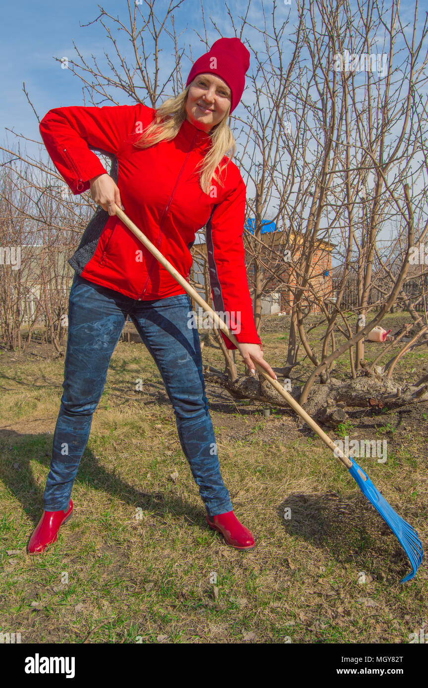 Junge schöne Frau Bauer reinigt Rechen trockenes Gras, das Tragen der roten Jacke, Stiefel und Hut, vertikale Foto Stockfoto