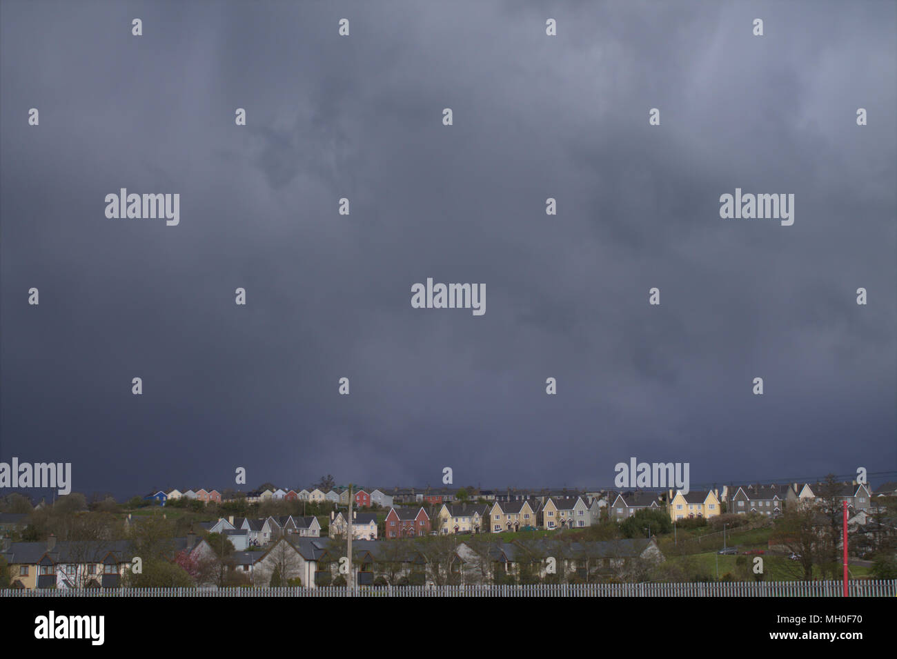 Schwere, dunkle Gewitterwolken über dem bunt bemalten Häuser auf dem Bergrücken oberhalb einer kleinen Stadt im County Cork, Irland. Stockfoto