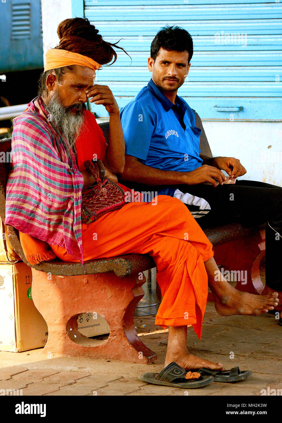 Zwei Männer in Indien sitzen auf einer Bank auf dem Bahnhof in Goa. Man im modernen Gewand, eine, vielleicht ein fakir, in traditioneller Kleidung. Margao, Goa Stockfoto