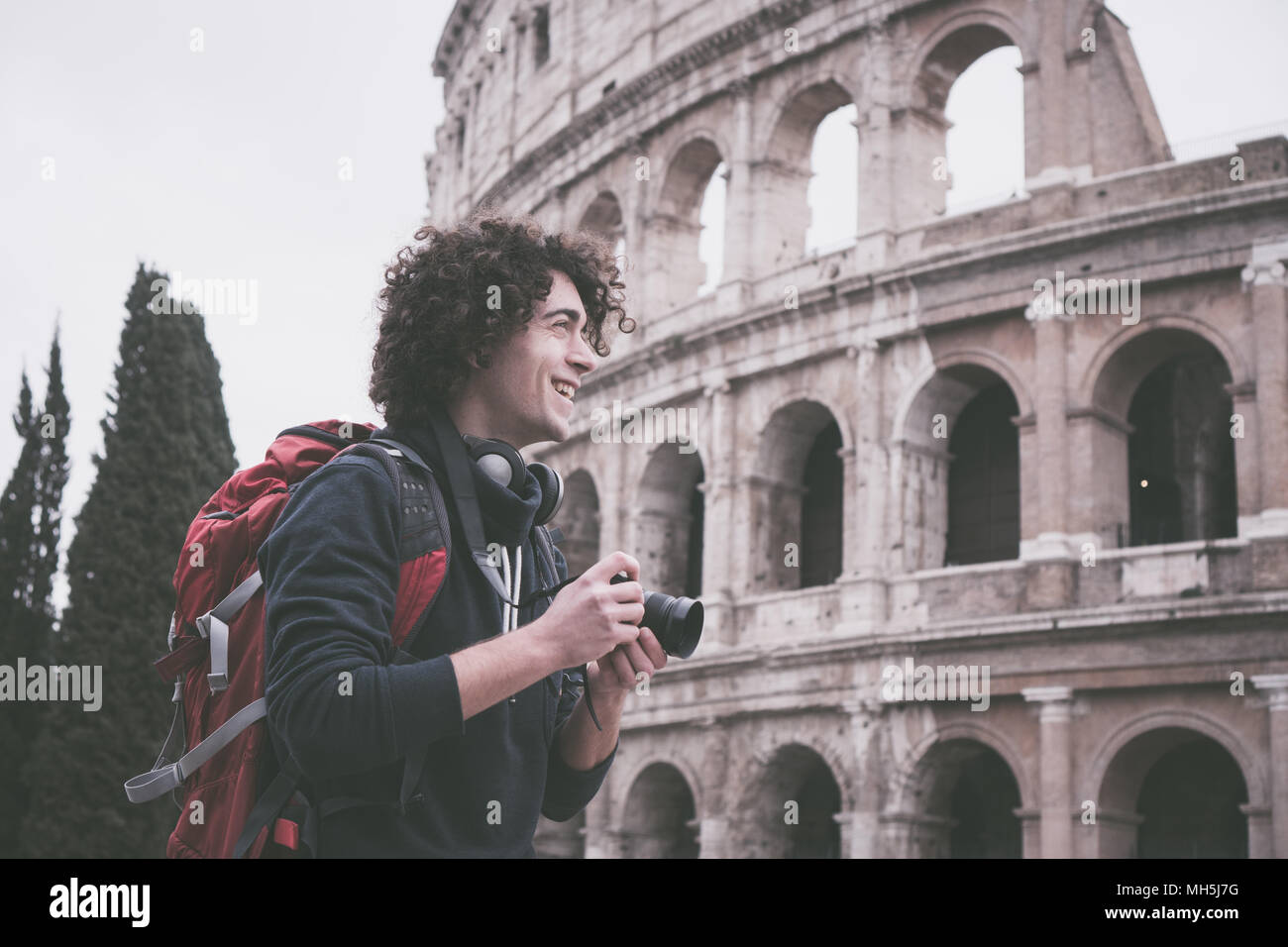 Hübscher junger Tourist Mann mit lockigem Haar mit Kamera und Rucksack, die Bilder von Kolosseum in Rom, Italien. Jahrgang suchen Image der jungen Tour Stockfoto