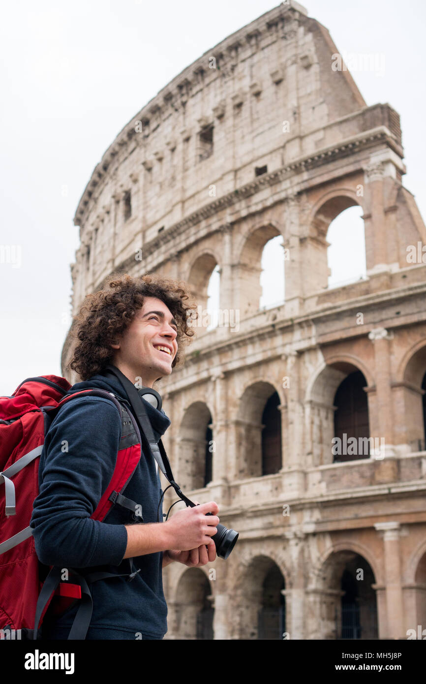 Hübscher junger Tourist Mann mit lockigem Haar mit Kamera und Rucksack, die Bilder von Kolosseum in Rom, Italien. Jungen Touristen, die Bilder von Co Stockfoto