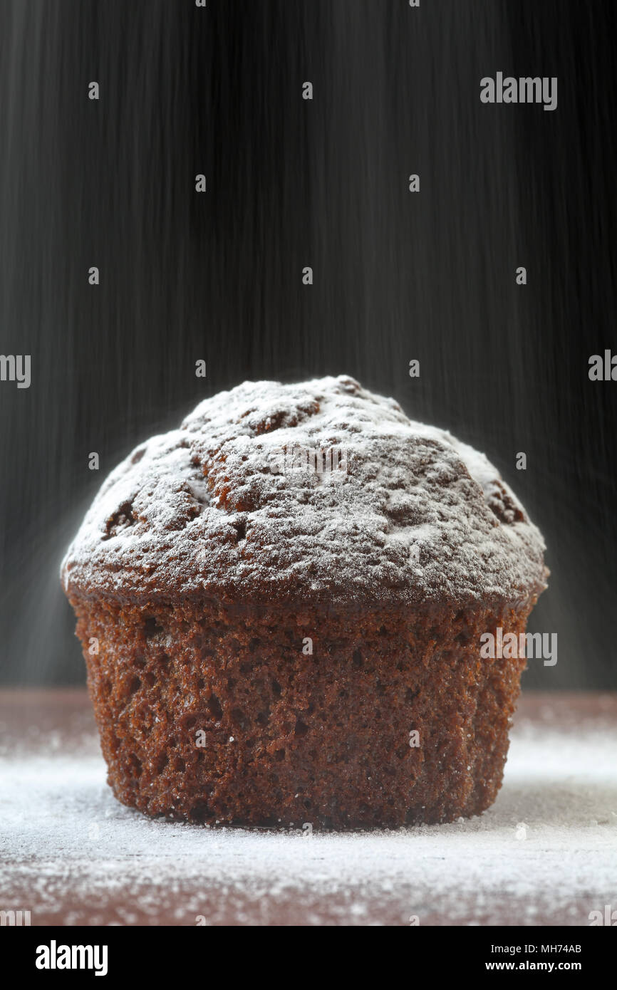 Nahaufnahme eines köstlichen Schokolade muffin auf einem Holztisch, auf schwarzem Hintergrund, mit Schnee wie Pulver Zucker um und über fallende verstreut. Stockfoto