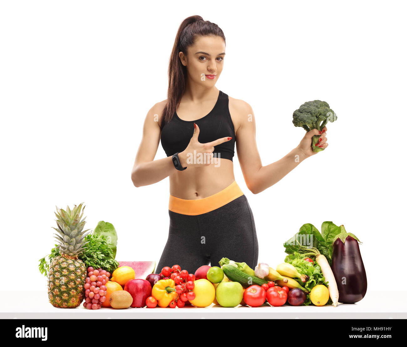 Fitness Frau hinter einem Tisch mit Obst und Gemüse, Brokkoli und zeigt auf weißem Hintergrund Stockfoto