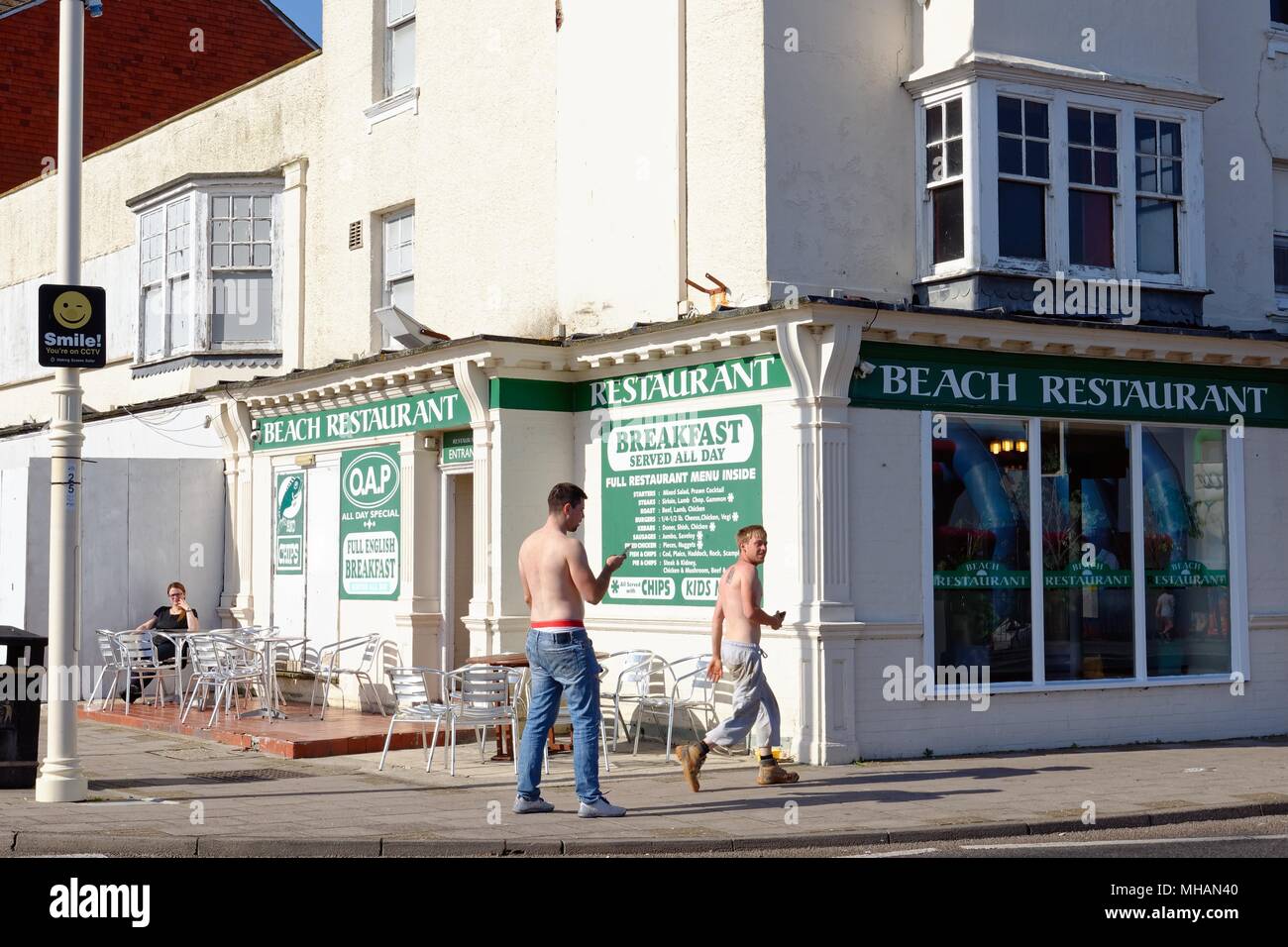 Zwei bare chested Jugendliche, eine auf Handy vorbei direkt am Meer Cafe mit Lady draußen zu sitzen, in Bognor Regis West Sussex England Großbritannien Stockfoto