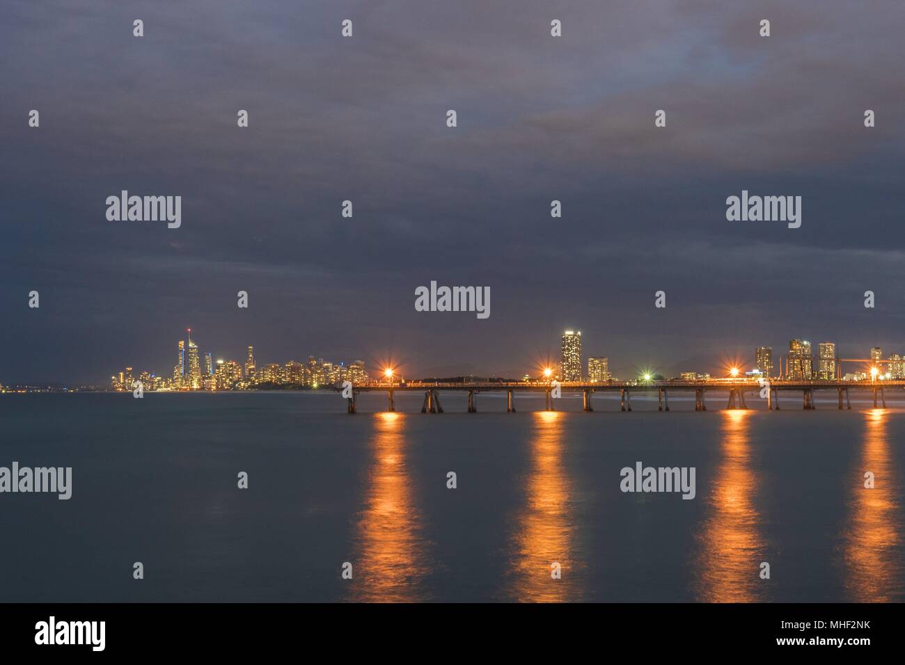 Die abenddämmerung Glühen von der Gold Coast reflektiert über das Wasser mit den Lampen mit einer Mole im Mittelgrund Lackierstraßen in den unscharfen Wasser. Stockfoto