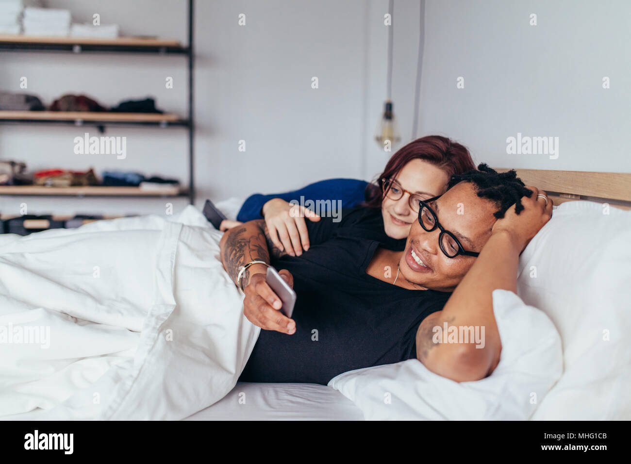 Glückliches Paar liegen auf einem Bett in einem Schlafzimmer und ein Handy. Gemischten rennen Mann und Frau im Bett mit Handy. Stockfoto