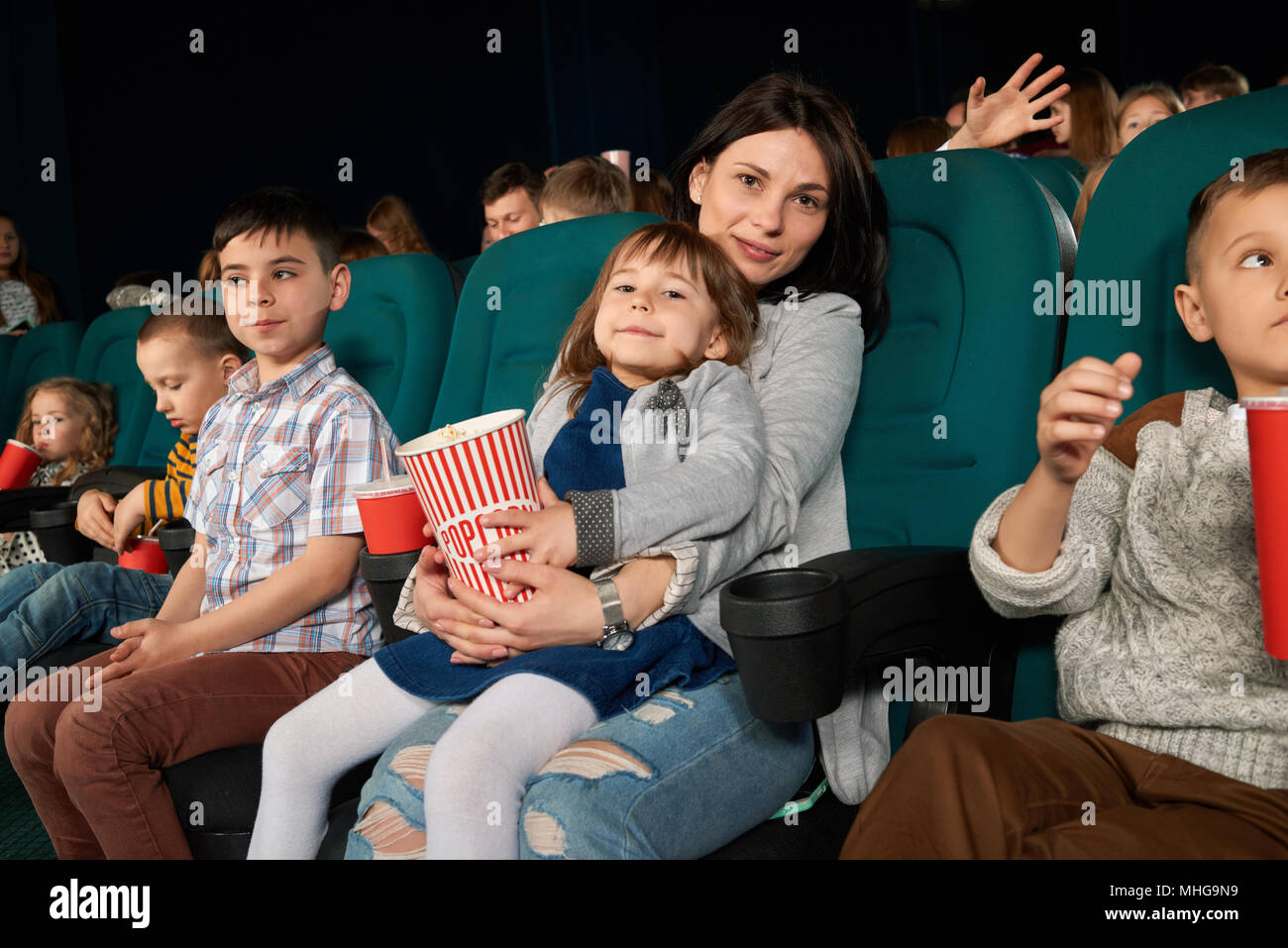 Zuschauer in bequemen Sesseln im Kino zu sitzen, aufzupassen Film. Schöne Frau und kleines Mädchen an der Kamera posieren, paperbag mit Popcorn in den Händen. Kinder trinken kohlensäurehaltige Getränke. Stockfoto
