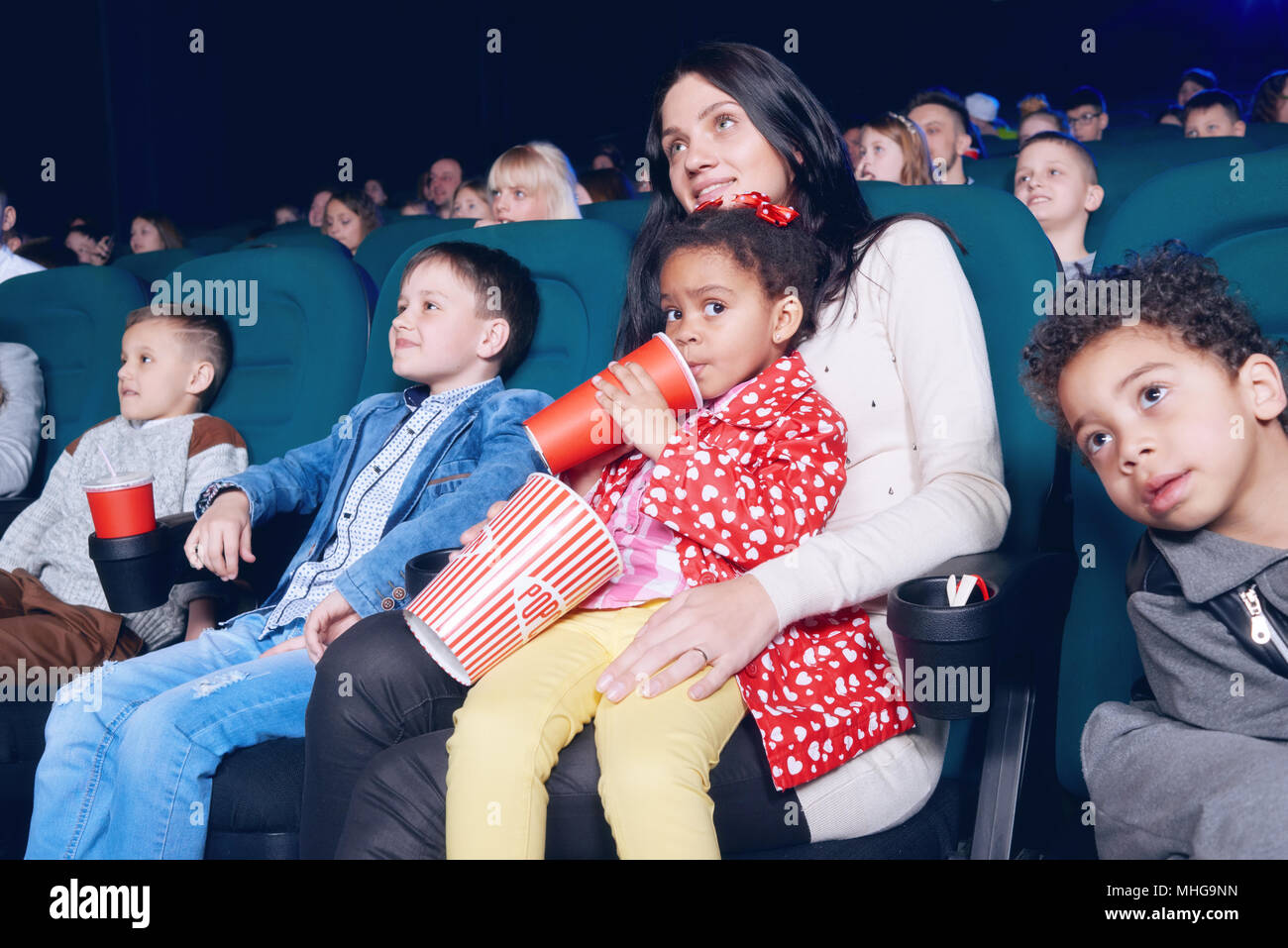 Schöne Frau und hübschen kleinen Mädchen im Kino posieren. Mädchen trinken fizzy Getränke und Popcorn Papiertüte. Kleine Zuschauer sitzen in bequemen Stühlen, gerade am Bildschirm und lächelnd. Stockfoto