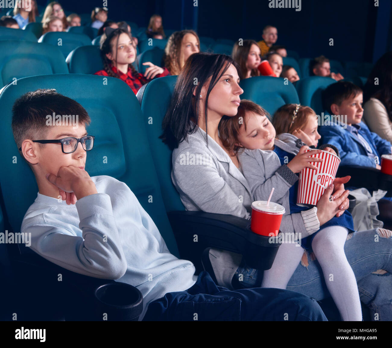 Die Zuschauer sitzen in bequemen Stühlen und beobachten Film im Kino. Junge schauen mit Interesse am Bildschirm mit unerschütterlichem Augen. Kleine hübsche Mädchen mit geschlossenen Augen sitzen auf ihrer Mama runden. Stockfoto