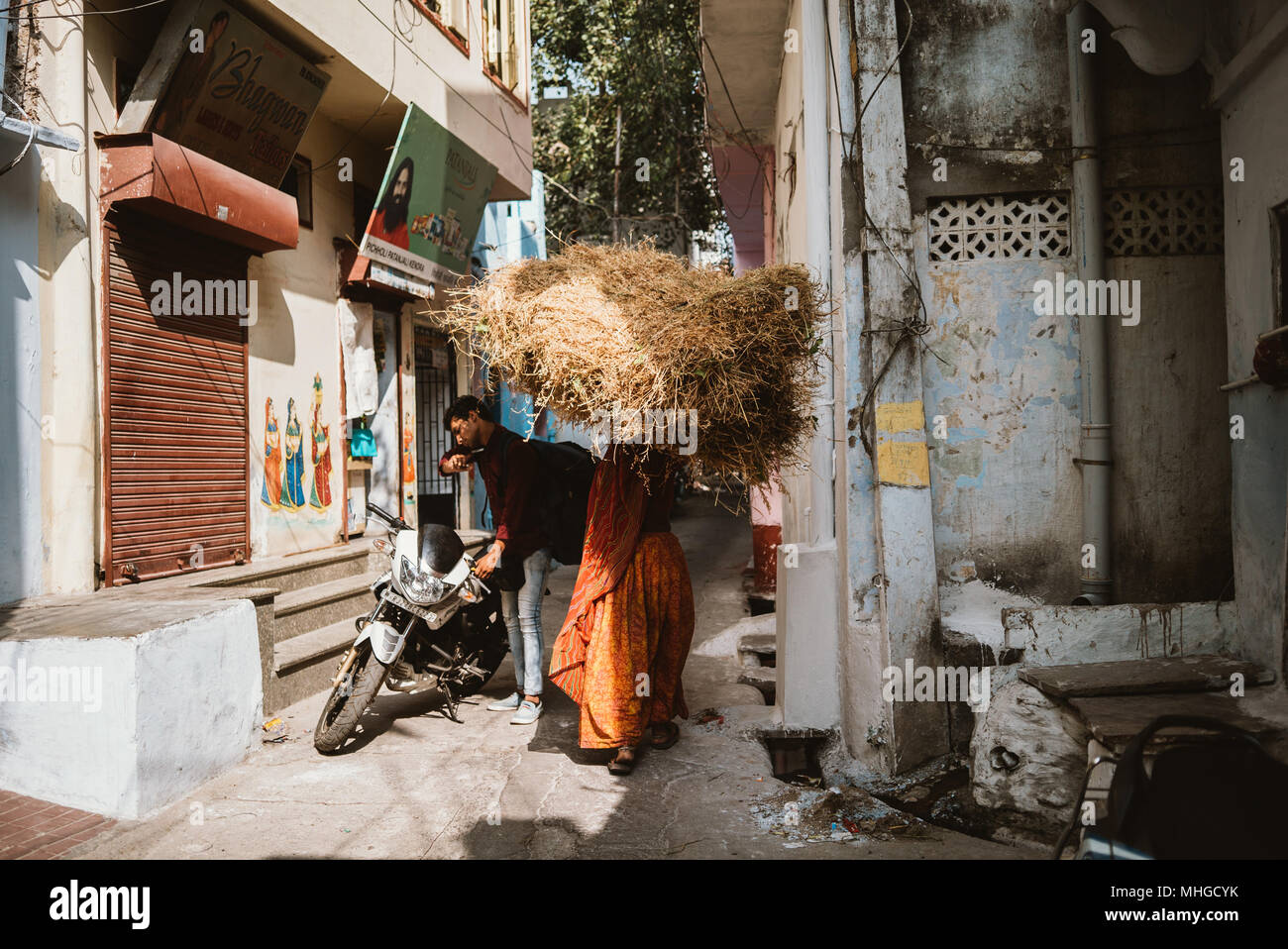 Eine Frauen Heu tragen auf dem Kopf geht ein Mann und sein Motorrad in einer schmalen Gasse in Udaipur, Indien Stockfoto