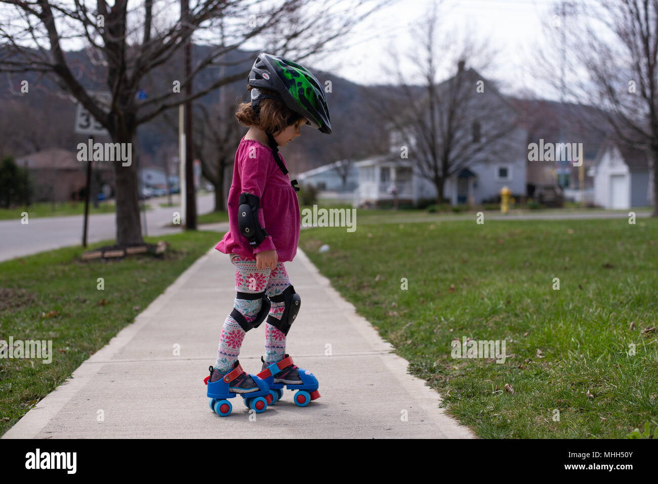Ein kleines Mädchen trägt einen Helm, Knie- und Ellenbogenschützer während Skaten auf einem Bürgersteig an einem sonnigen Frühlingstag. Stockfoto