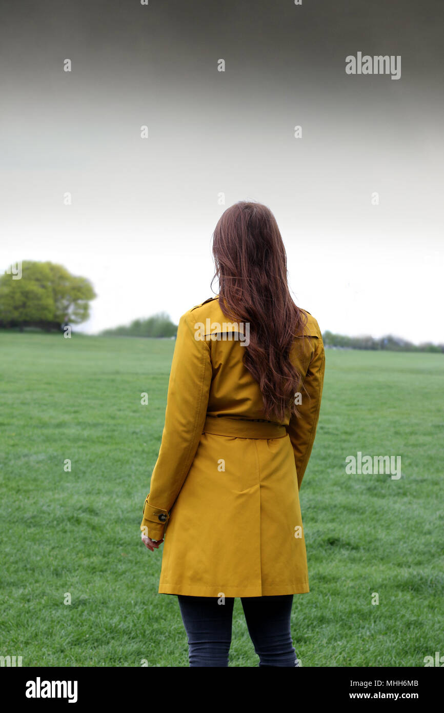 Junge Frau, die in einem Park mit dem Rücken zur Kamera mit Blick auf den Horizont, sie trägt ein Ocker Regenmantel. Stockfoto