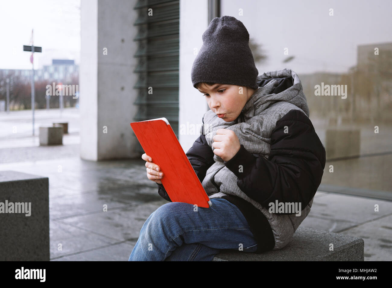 Aufgeregten kleinen Jungen spielen online Spiel auf Tablet Computer während draußen sitzen nur auf der Straße im Winter feiern gewinnen mit geballten Faust Stockfoto