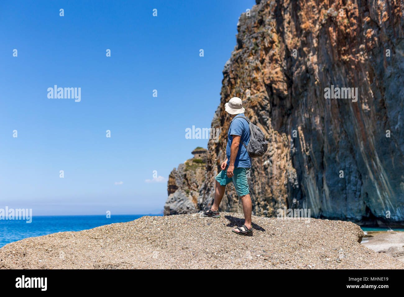 Sommer reisen Konzept. Rückansicht Blickrichtung Porträt einer männlichen Backpacker im Sommer Outfit am wunderschönen Strand von Felsen umgeben. Horizontale erschossen. Platz kopieren Stockfoto