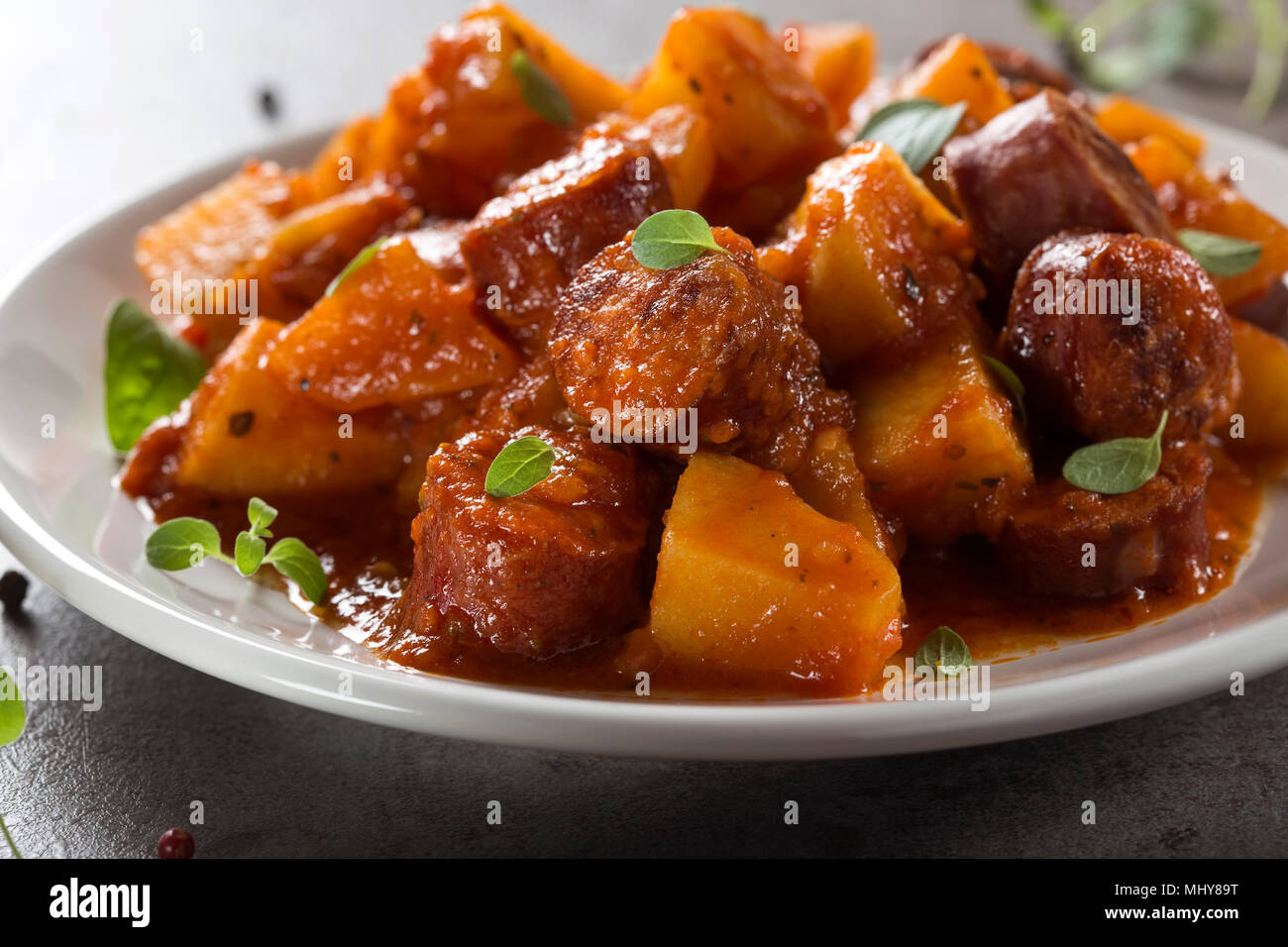 Kartoffeln Eintopf mit Schweinefleisch Wurstscheiben und Tomatensauce auf einem weißen Teller mit Oregano Blätter Stockfoto