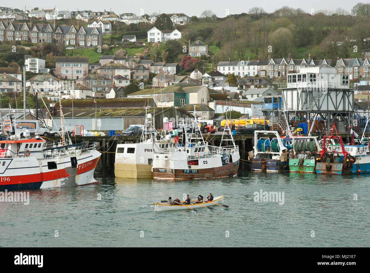 Die traditionelle Cornish racing gig Pendeen Ausbildung in Newlyn Harbour in den Vordergrund mit Fischereifahrzeugen und die Stadt von Newlyn im Hintergrund. Stockfoto