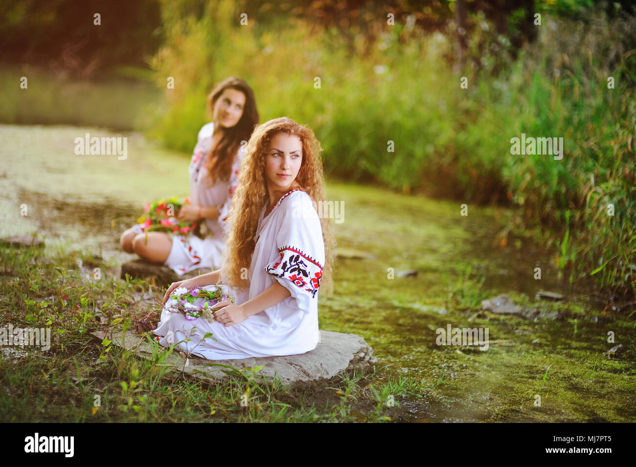 Zwei junge schöne Mädchen in weißem Hemd mit floralen Ornament mit Kränze in ihren Händen sitzen auf dem Hintergrund des Flusses. Stockfoto