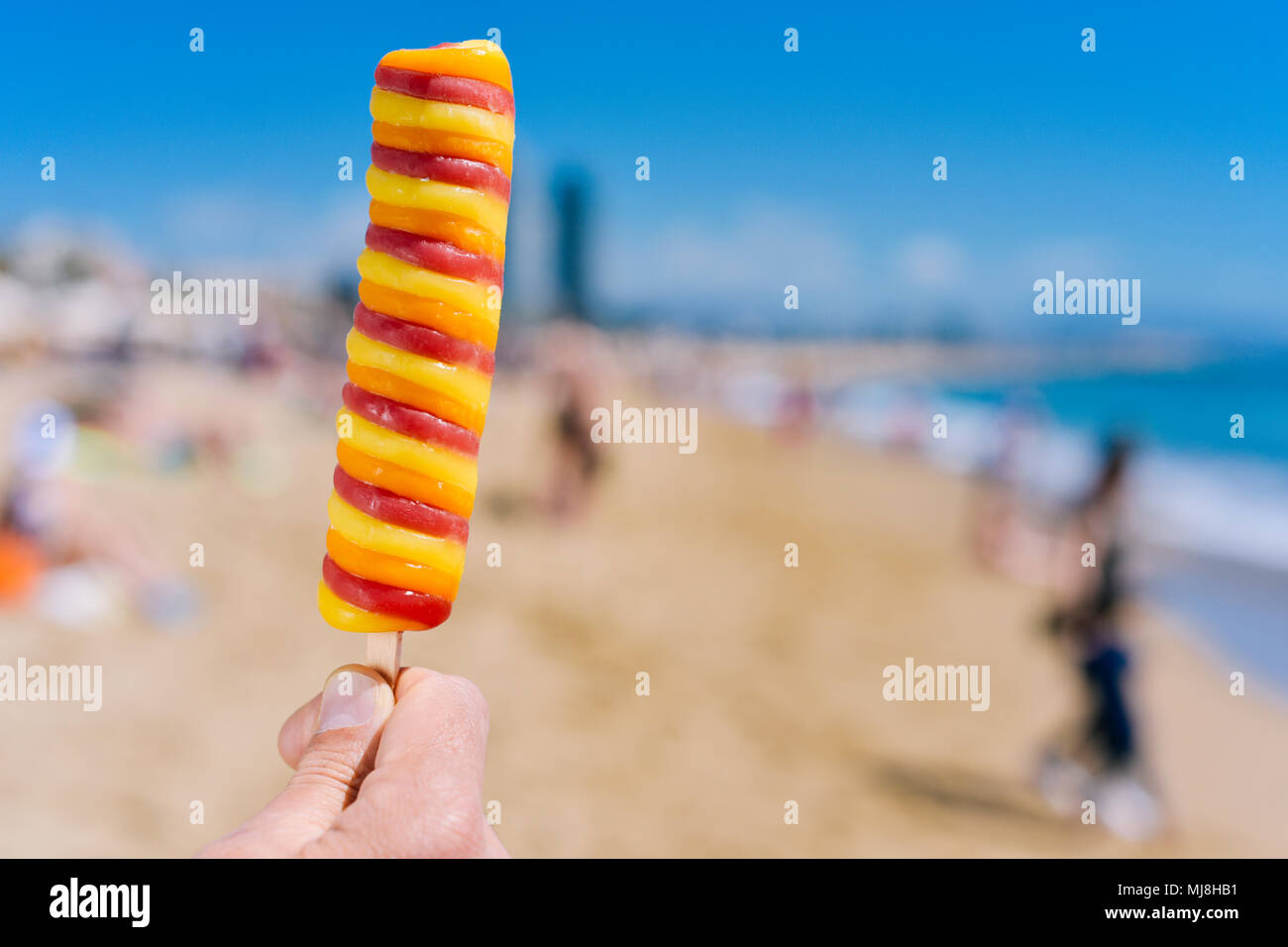Nahaufnahme von einem erfrischenden Popsicle in der Hand eines jungen kaukasischen Mann am Strand, mit unkenntlich Leute im Hintergrund Stockfoto