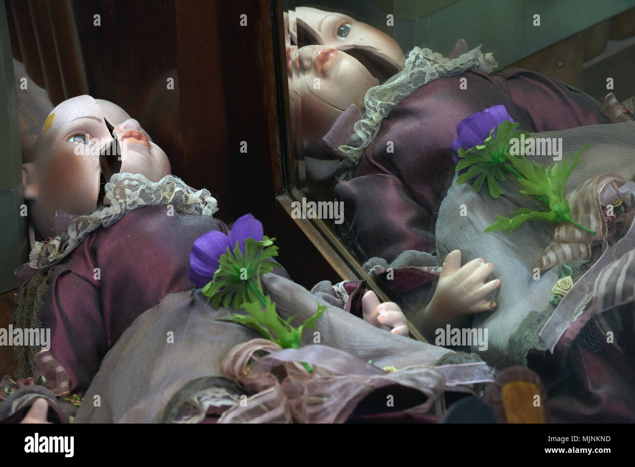 Alte Porzellanpuppe mit einem gebrochenen Kopf in ein altes Kleid in der Nähe der alten Spiegel spiegeln sich darin, ein Foto von einem modernen Design der Wanddekoration. Stockfoto