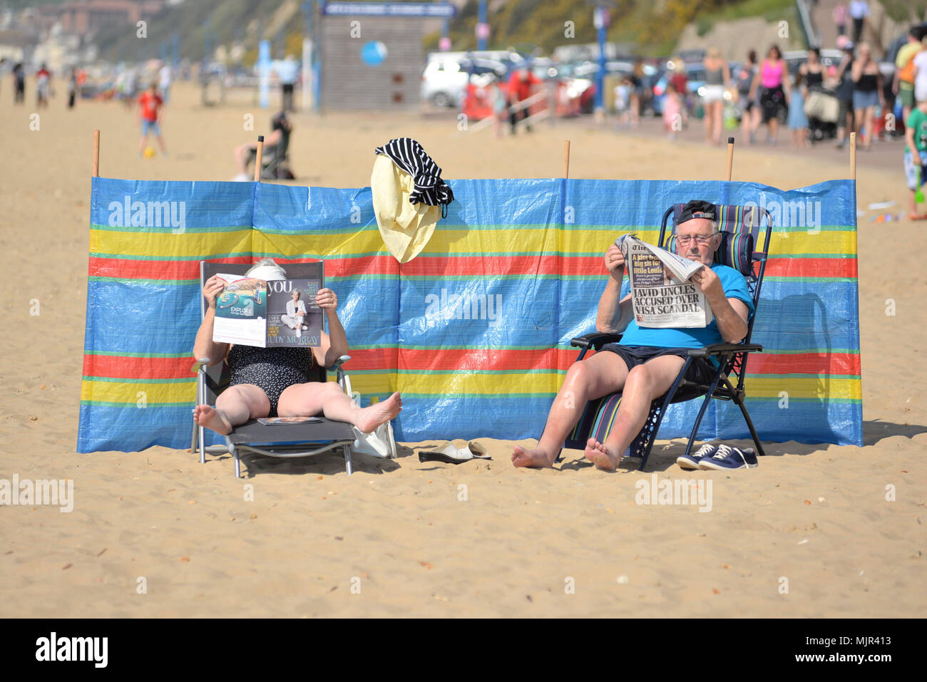 Boscombe, Bournemouth, Dorset, Großbritannien, 6.. Mai 2018: Paar sitzt auf Stühlen und liest Zeitungen und entspannt sich am Strand bei heißem Sonnenschein, geschützt durch eine Windpause. Heißeste Mayday-Feiertagswochenende auf Rekordzeit. Stockfoto