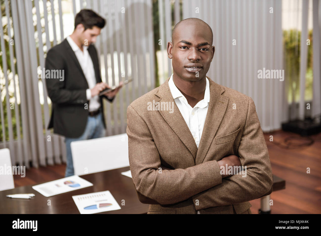 Zuversichtlich Geschäftsmann. Fröhlicher junger afrikanischer Mann in formale halten die Arme verschränkt und während seine kaukasischen Kollegen stehen auf Hintergrund lächelnd Stockfoto