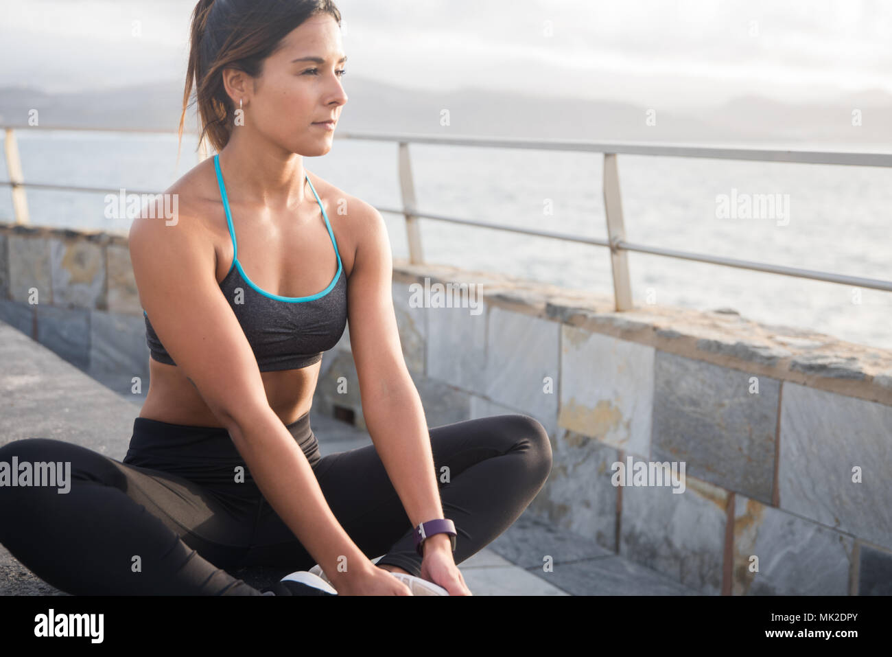 Hübsche junge sportliche Dame in Active Wear sitzt mit den Beinen auf den Boden und die Füße zusammen, während sie auf das Meer Stockfoto