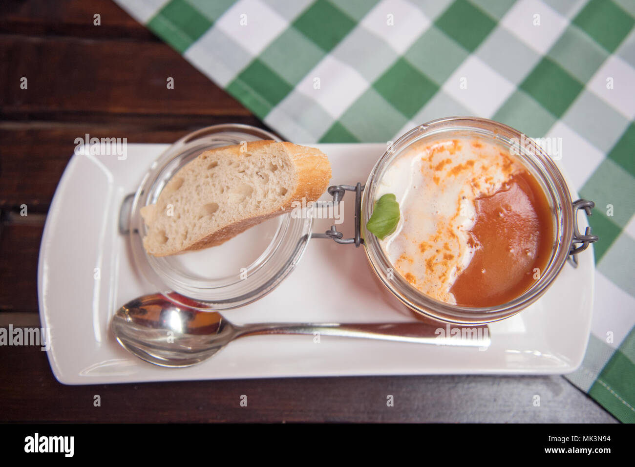 Eine köstliche Portion cremiger Tomatensuppe in einem Glas, garniert mit Basilikum, begleitet von einem Stück frischem Brot auf einem weißen Teller, Stockfoto