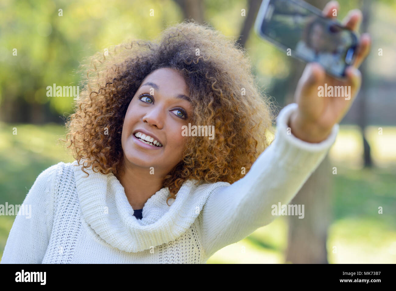 Schöne junge afrikanische amerikanische Frau mit Afro Frisur und grüne Augen tragen weiße Winter Dress. Schöne Mädchen selfie im Park mit einem Smartph Stockfoto