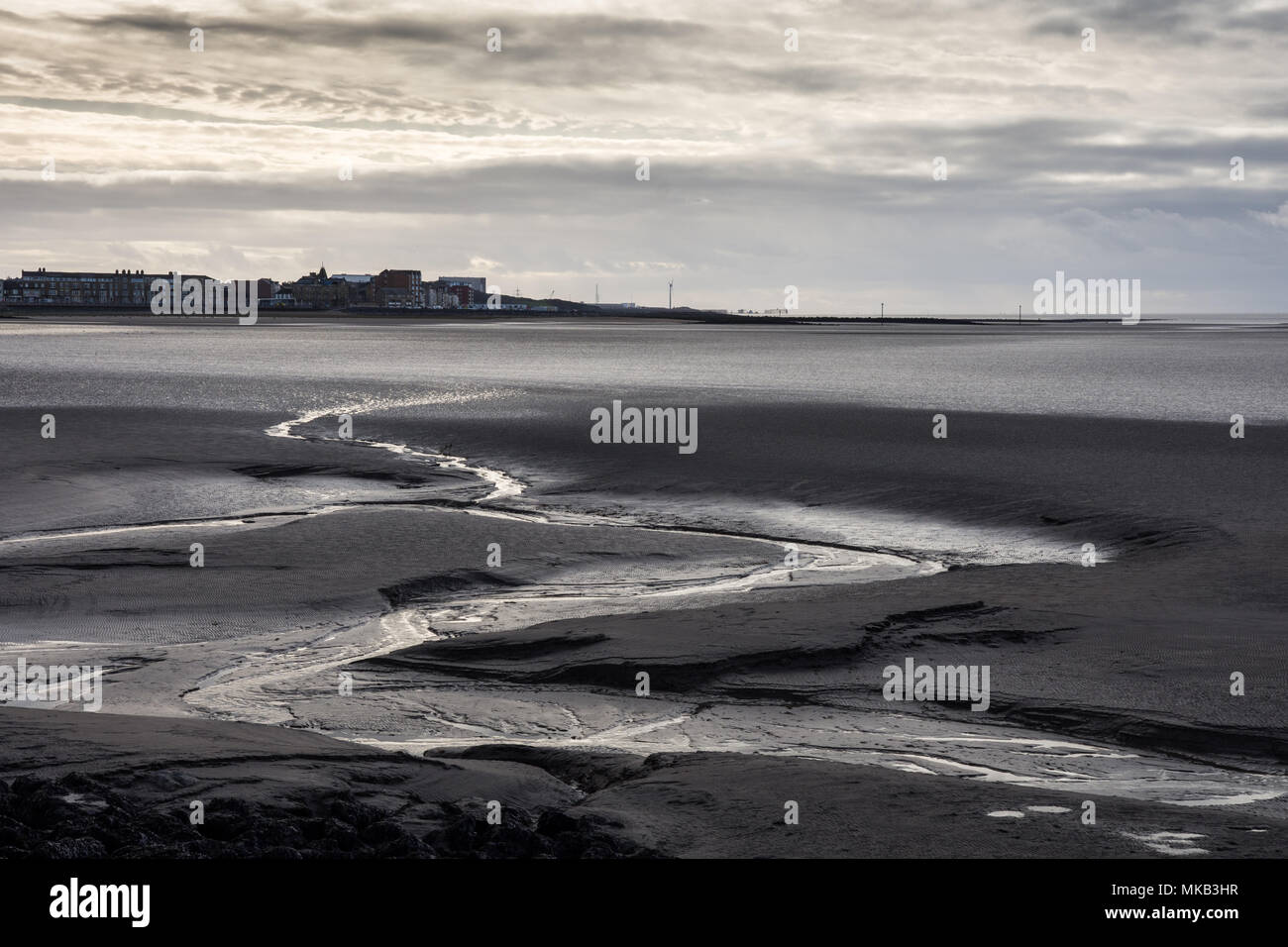 Ein Bach schlängelt sich durch das Watt bei Ebbe in Morecambe Bay, mit der Strandpromenade von sandylands und Heysham Power Station im Hintergrund. Stockfoto