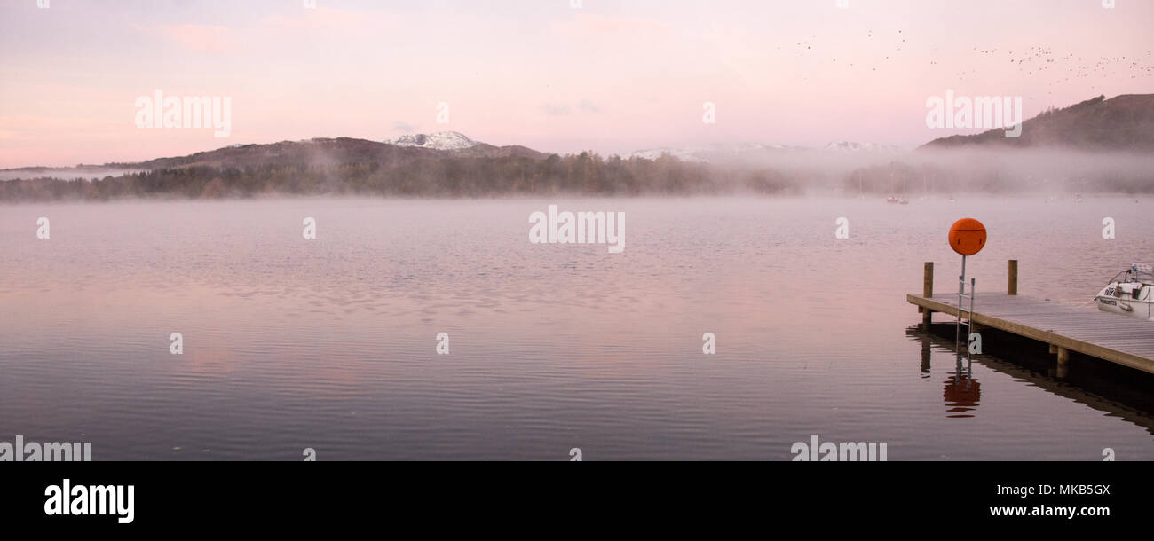 Nebel steigt aus den ruhigen Gewässern des Windermere Lake in Ambleside Pier neben Herbst Wald und unter Thew schneebedeckten Bergen des Langdale, in Engl Stockfoto