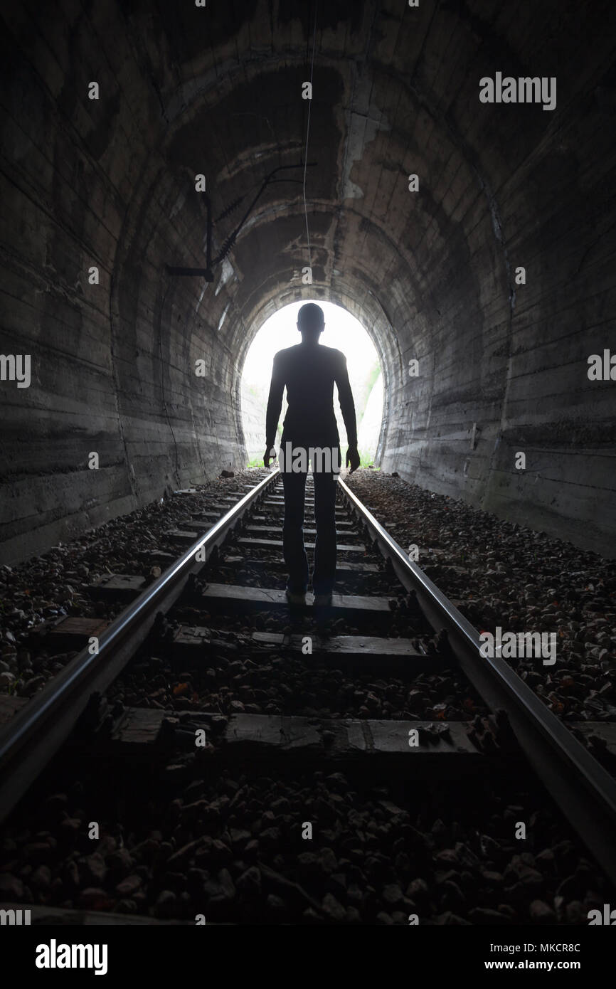 Mann Silhouette, die in einem Tunnel in der Mitte der Gleise mit Blick auf das Licht am Ende des Tunnels in einem konzeptionellen Bild Stockfoto