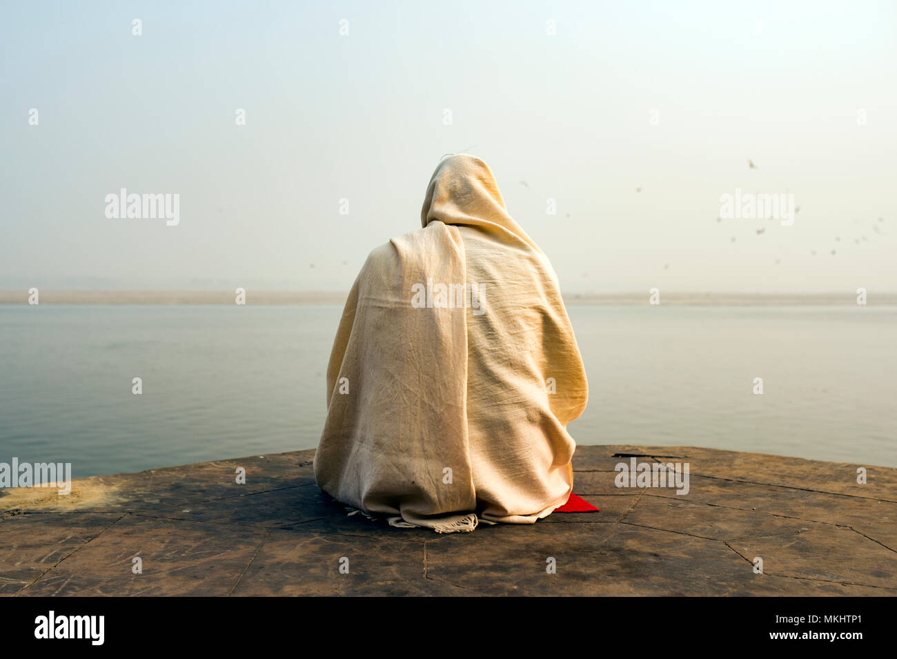 Ein heiliger Mann in Weiß gekleidet ist betend und meditierend auf einem der vielen Ghats von Varanasi vor dem heiligen Fluss Ganges in Indien. Stockfoto