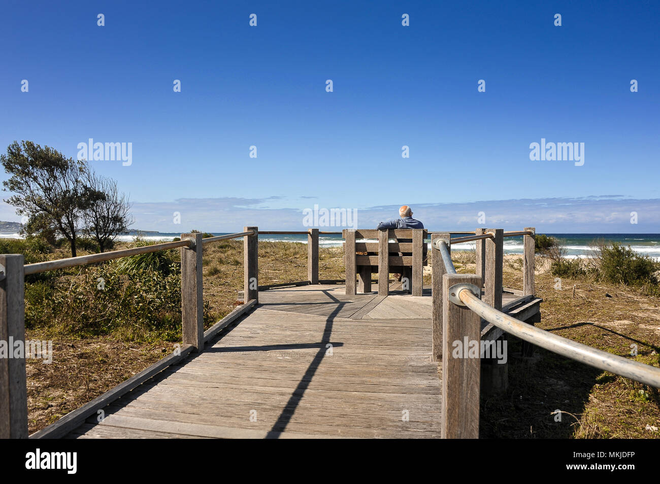 Man entspannt sich auf Holzbank, beobachten die Wellen entlang Seven Mile Beach, Australien. Küsten Szene, Holzsteg, grasbewachsenen Dünen und blauer Himmel Stockfoto