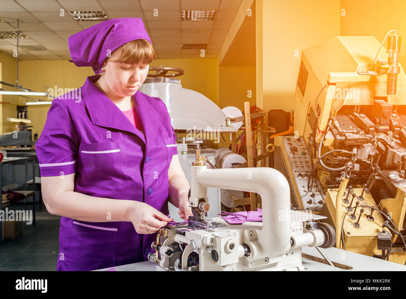 Eine junge Frau arbeitet als Näherin in einer lila Unifrome ist echt Leder Kinder Schuhe auf eine Nähmaschine näht, im Hintergrund eine Nähmaschine arbeiten Stockfoto