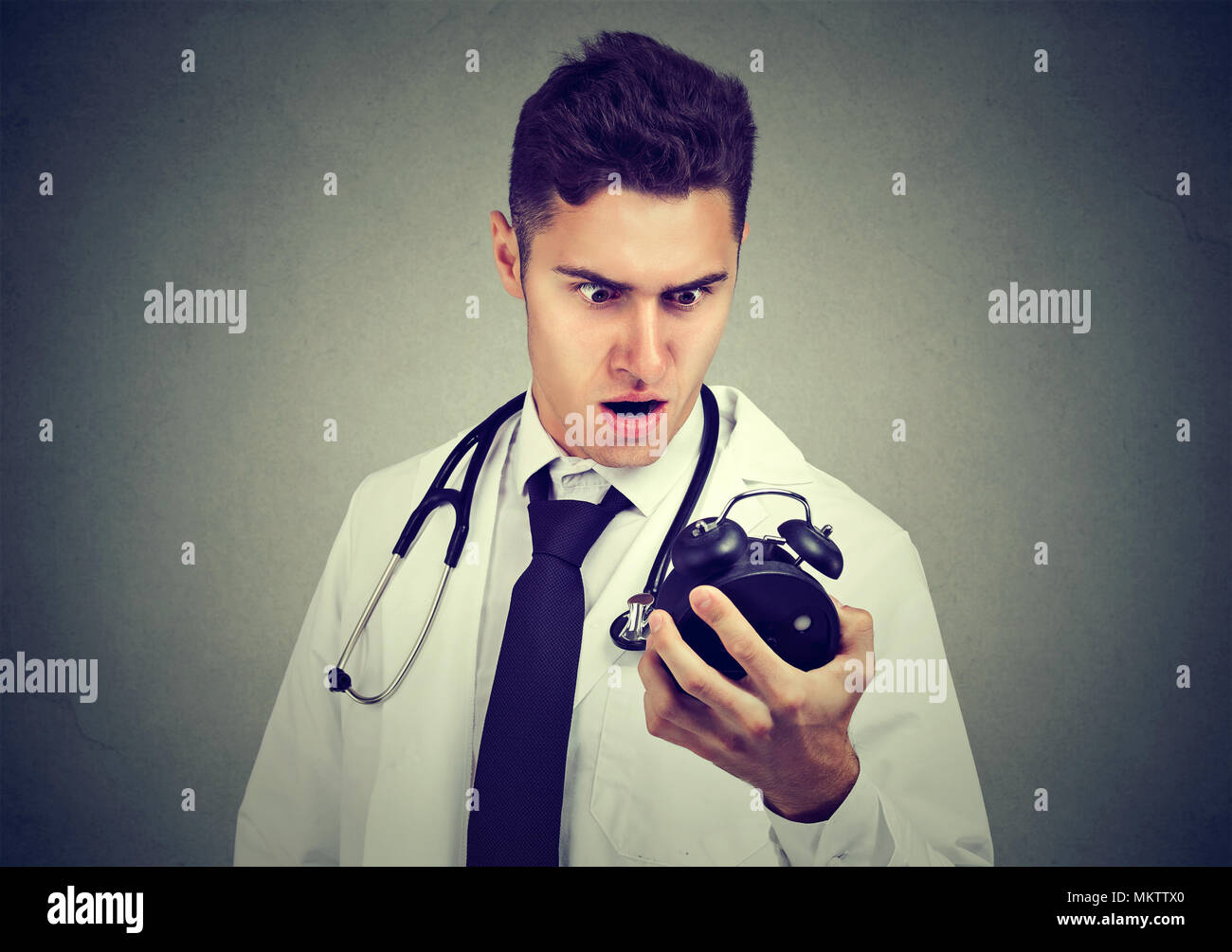 Junge Mann in der medizinischen Outfit mit der Zeit Druck auf die Uhr auf grauem Hintergrund suchen Angst. Stockfoto