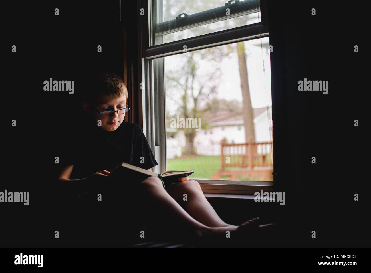 Ein 11-jähriger Junge sitzt in einem offenen Fenster und liest ein Buch. Stockfoto