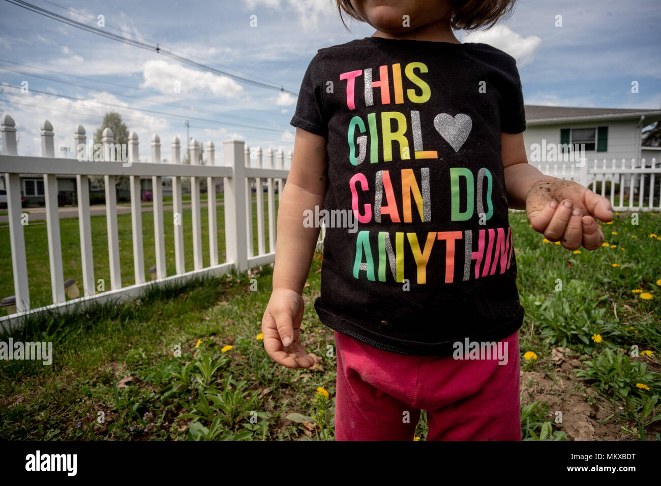 Ein kleines Mädchen, ohne ihr Gesicht zu sehen ist, trägt ein T-Shirt in einem Garten mit einem weißen Zaun hinter ihr. Stockfoto