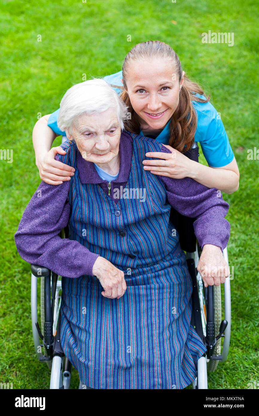 Portrait von ältere behinderte Frau in einem Rollstuhl verbringen Zeit im Freien mit fröhlichen jungen Betreuer Sitzung Stockfoto
