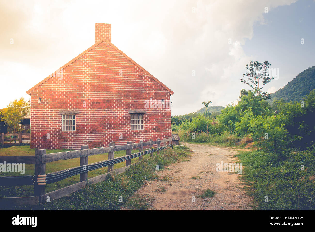 Wunderschöne Landschaft Bild des alten Backsteinhaus mit hölzernen Zaun neben Dirt Road an der Landschaft im regnerischen Tag umgeben. Stockfoto