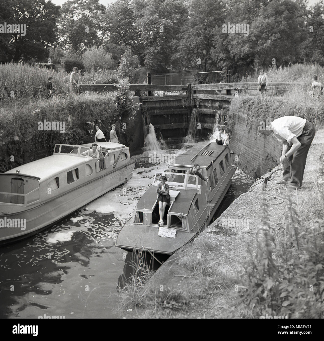 1960, historische, zwei schmale Boote nebeneinander in eine Schleuse, die Schleusen geöffnet, England, UK. Diese hölzernen Tore oder Schranken steuern die Wasserstände in den Sperren und damit anheben oder die Boote auf einer anderen Ebene zwischen erstreckt sich von Wasser auf den Kanal oder Wasserstraße. Die Schleuse wurde von Leonaro da Vinci erfunden. Stockfoto
