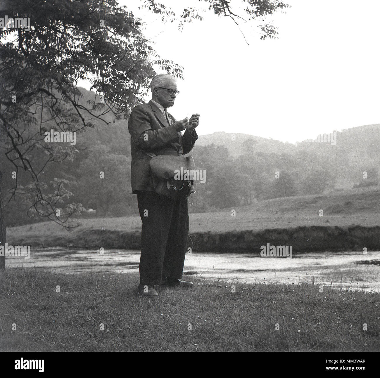 1950 s, historischen, alten Mann auf Ufer mit Leder Tasche oder Flaschenzug Beutel, seinen Angriff bereit für Fliegenfischen, England, UK. Stockfoto