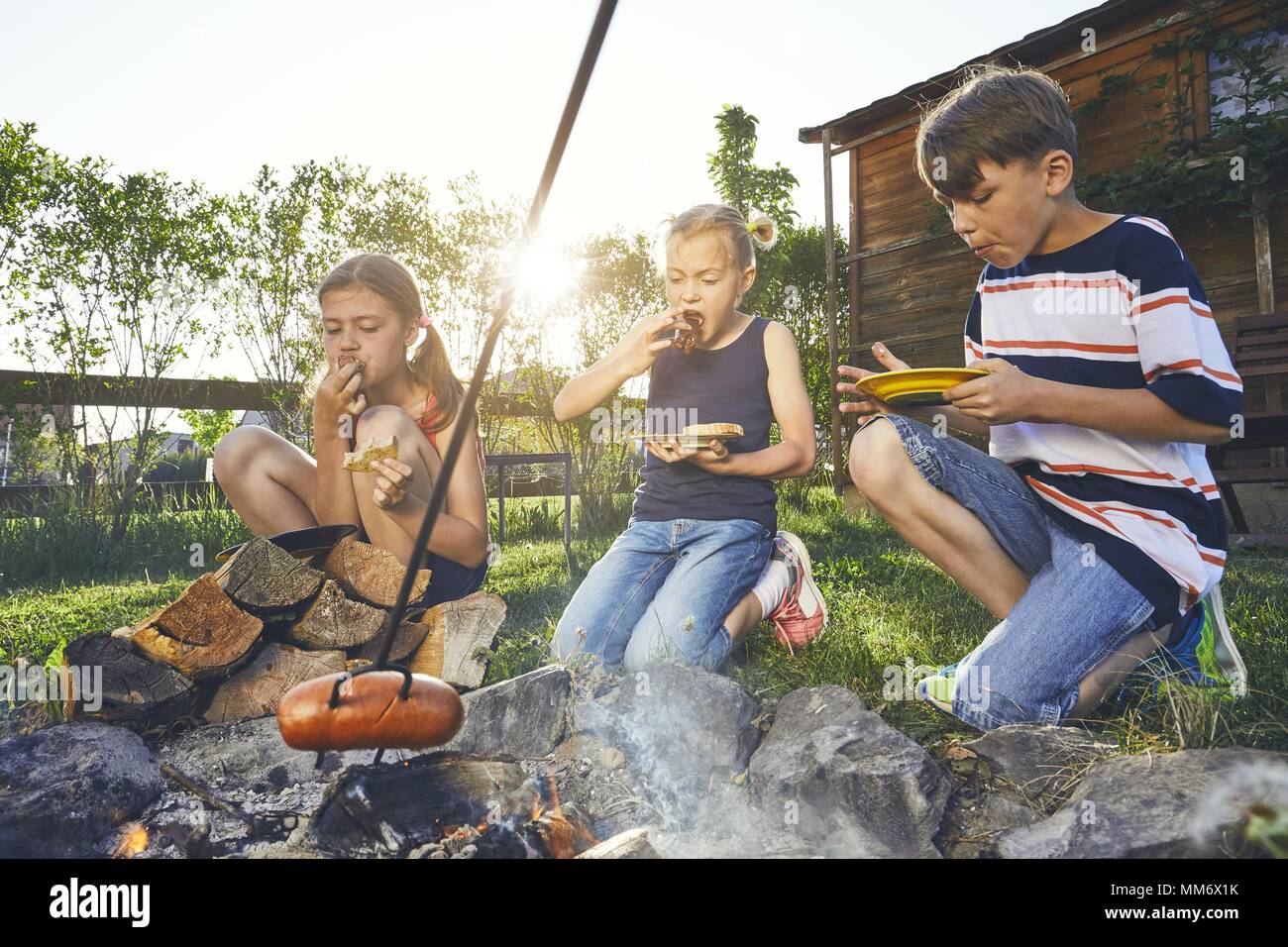 Kinder genießen, Lagerfeuer. Geschwister (Familie) toasten Würstchen auf den Garten bei Sonnenuntergang. Stockfoto