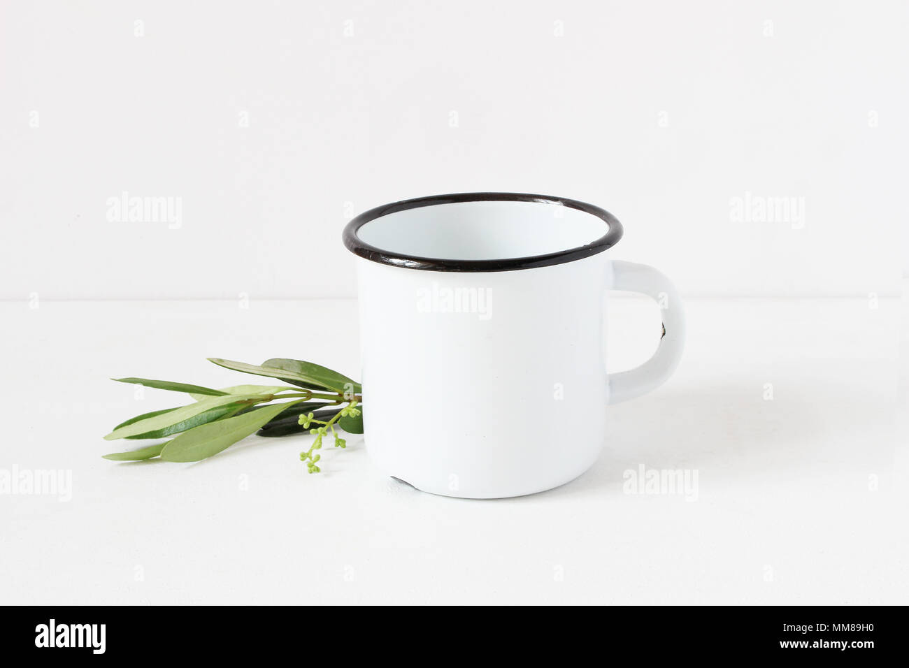Styled Foto. Feminine Stilleben Komposition mit leeren weißen Metall/Emaille Tasse und grüne Olive Branch auf weißem Hintergrund. Sommer rustikalen Szene, Produkt mockup. Stockfoto