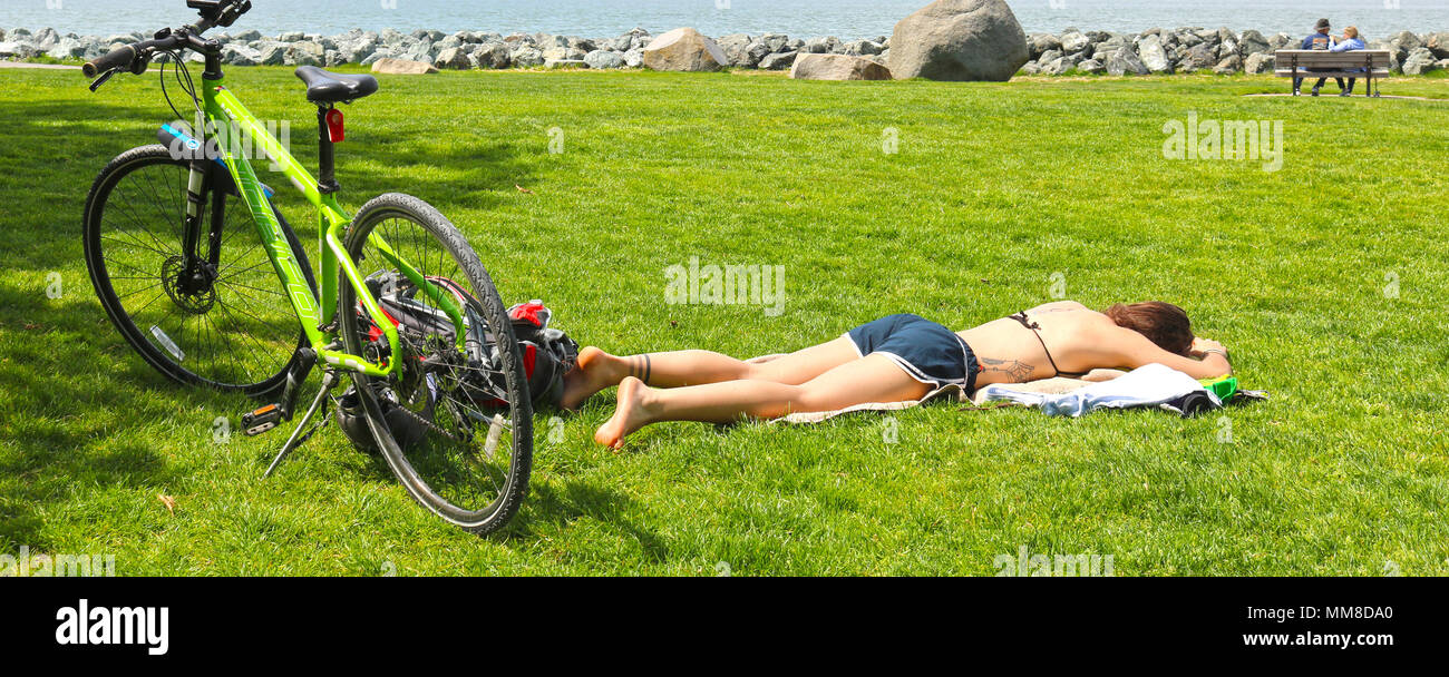 Junge Frau mit in das Gras am Boulevard Park Liegewiese, während ihr Fahrrad hinter ihr ist. Ein älteres Paar auf einer Parkbank in der backgrund sitzt Stockfoto