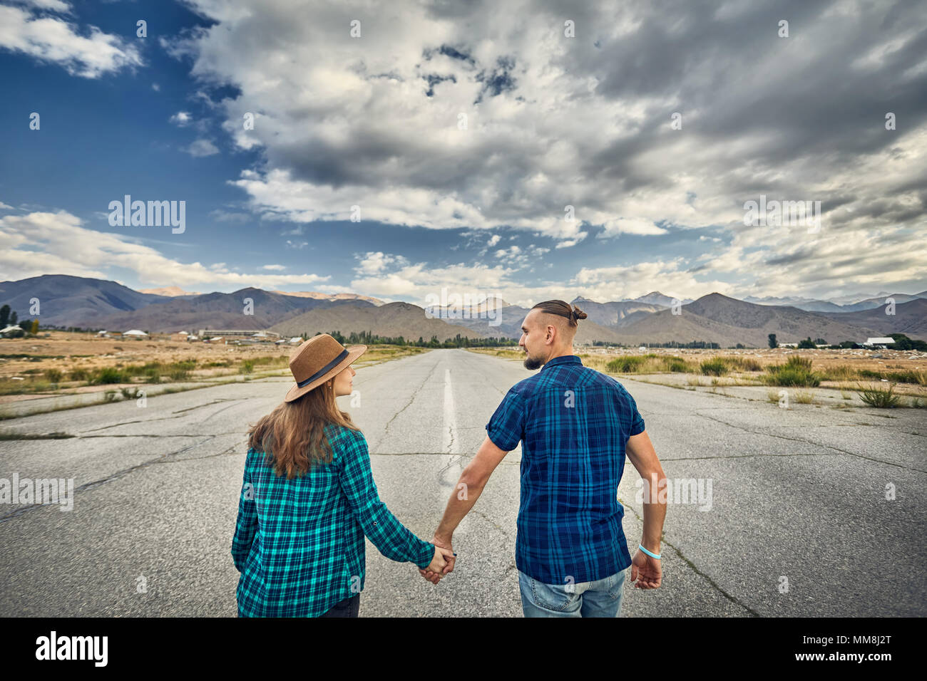 Glückliches Paar in kariertes Hemd Holding durch die Hände und laufen auf dem breiten Asphaltstraße mit Bergen und bewölkter Himmel Hintergrund Stockfoto