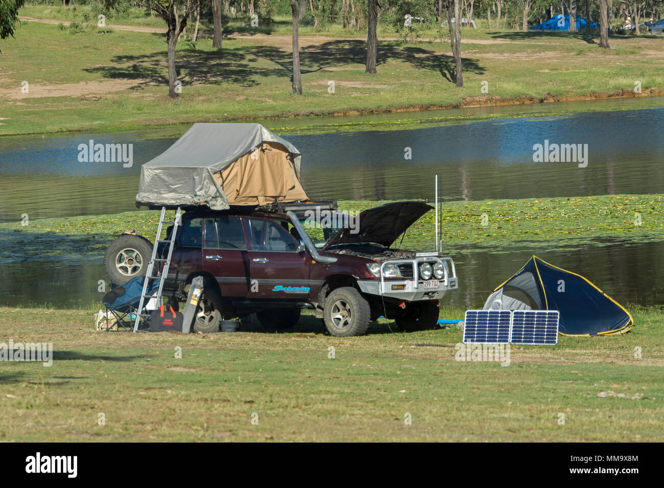 Fahrzeug mit Allradantrieb auf dem Dach Zelt, Campingausrüstung, und solar panel auf grassy Bank neben ruhigen blauen Wasser des Eungalla Dam, QLD, Australien Stockfoto