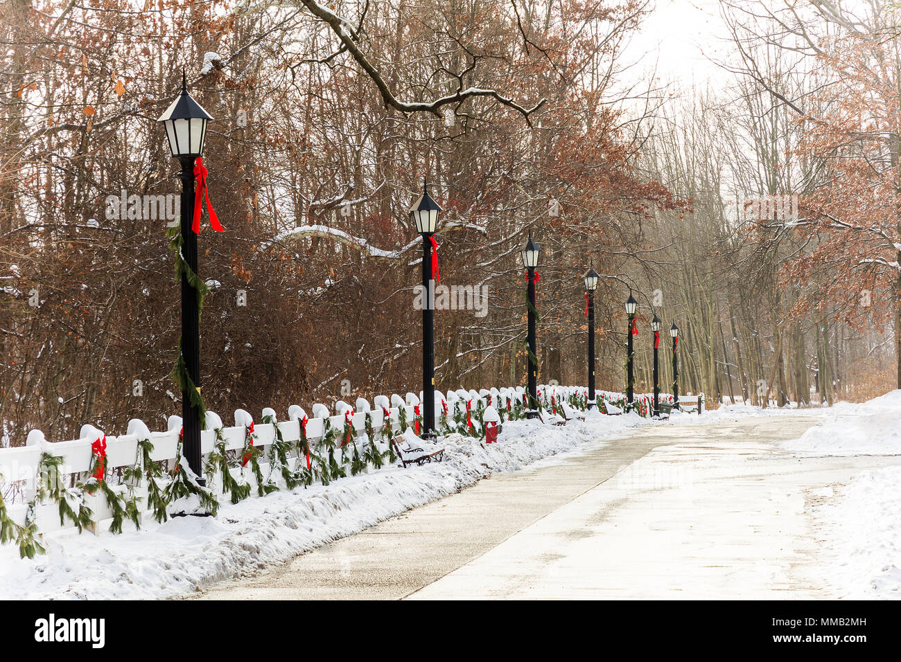 Eine verschneite Straße, gesäumt von alten Stil lampe Beiträge und einem weißen Holzzaun, mit immergrünen Kranz und roten Bändern geschmückt wurde. Stockfoto