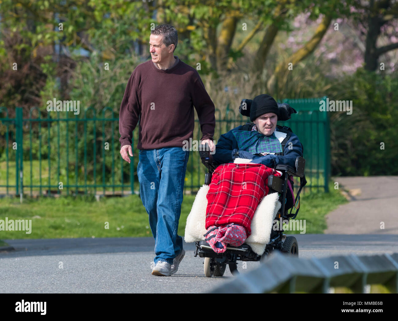Männliche Pflegeperson helfen einem Menschen mit offensichtlichen Behinderungen im Rollstuhl, wandern durch einen Park in Großbritannien. Stockfoto
