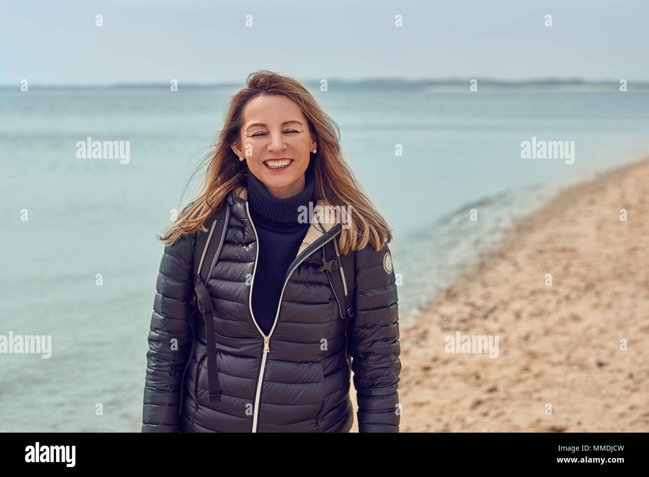 Attraktive Frau zu Fuß am Strand an einem bewölkten Tag trägt einen Rucksack und Jacke anhalten am Rande des Meeres zu lächeln in die Kamera Stockfoto