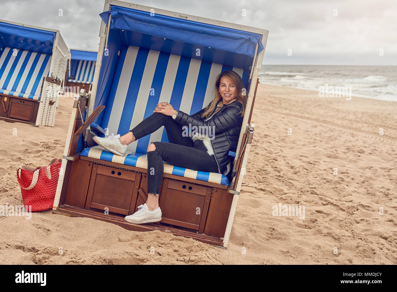 Attraktive schlanke blonde Frau sitzt in einer Strandhütte mit blau und weiß gestreifte Textil an einem Sandstrand an einem bewölkten Tag in die Kamera lächeln Stockfoto